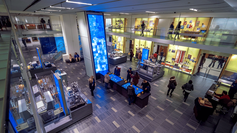 Visitors exploring the Interactive Media Column at the Science History Institute with several guests engaging with the touchscreen display while others walk through the exhibit space.