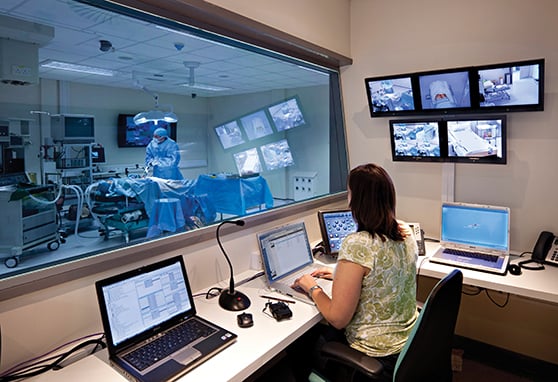 A medical control room viewed through a large observation window into an operating theater, where a surgeon in blue scrubs performs a fake procedure on dummy. In the control room, a technician sits at a desk with three laptops, a touch-panel console, a microphone and multiple wall-mounted monitors displaying live video feeds of the training exercise surgery.