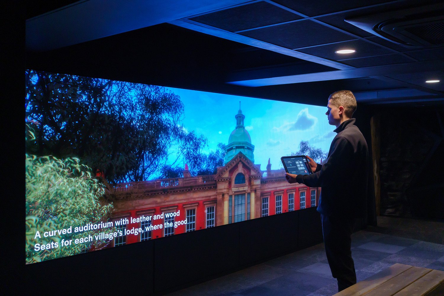 A man with a tablet looking at a video wall that displays the Miners Hall.