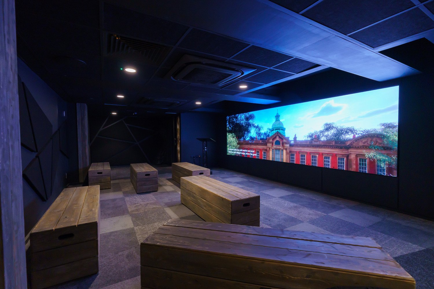 A black room with 6 wooden crates as furniture and video wall that displays media.