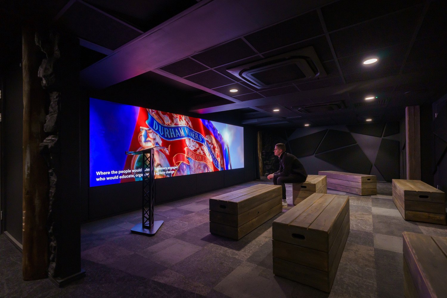 A man sitting on a wooden crate looking at the media on the wall with the Durham Miners Association on it.