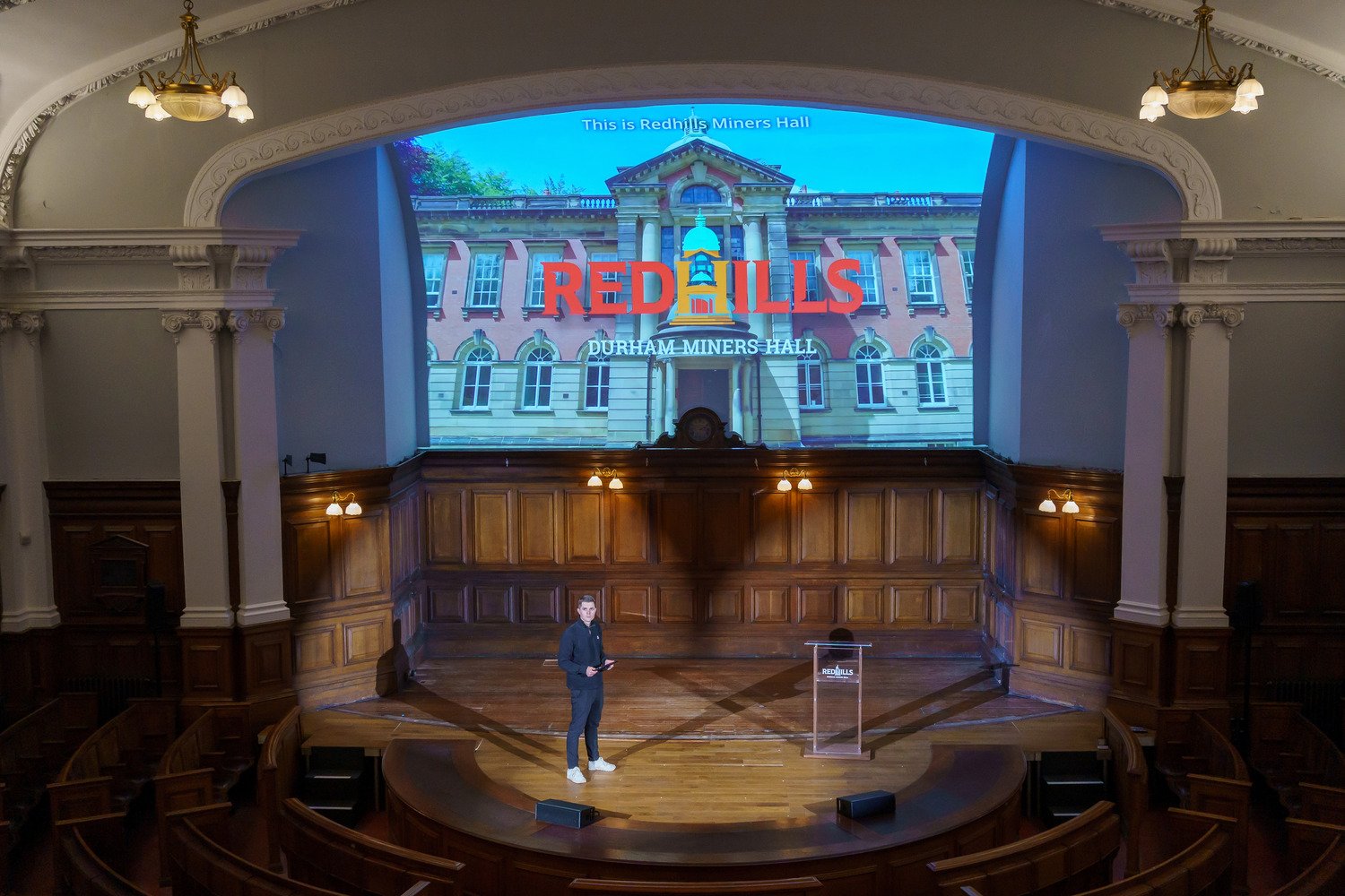 A man on the stage in the Pitman's Parliament with media displayed on the background.