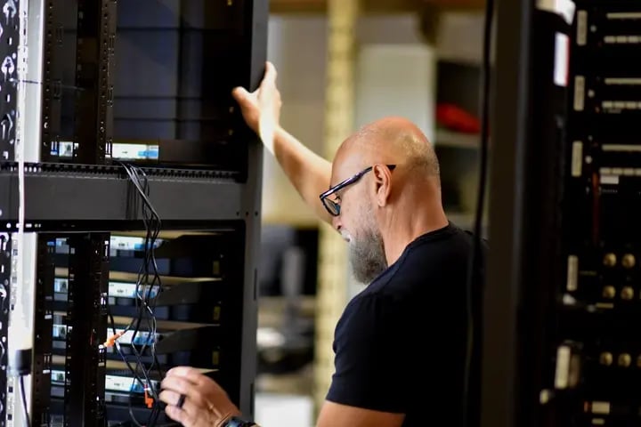 A man wearing a black shirt is engaged in server maintenance in an audiovisual rack room center.