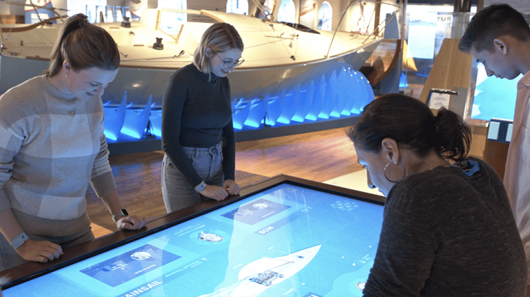 Four visitors engaging with an interactive touch table at The Sailing Museum, exploring digital exhibits in a modern, hands-on gallery space.