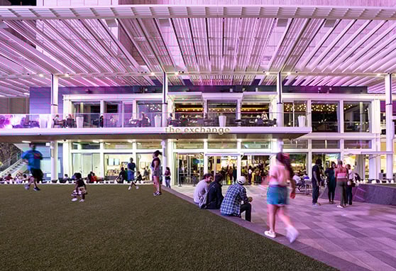 The Exchange, a multi-level dining and social hub. The setting exudes energy and modernity, emphasized by the dynamic lighting in soft purples and pinks, reflecting off the sleek metal and glass façade. The open-air structure and transparent design make it feel welcoming and connected to the surrounding plaza.