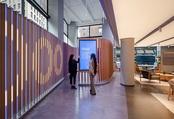 This Okta space blends modern tech with warm, natural design elements, featuring a dynamic digital display embedded within a curved wooden slat wall and a cozy seating area that encourages interaction and comfort.