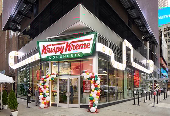 This vibrant Krispy Kreme storefront stands out with its bold signage, retro-style lighting, and festive balloon arches, creating an inviting atmosphere on the busy street corner. The glass exterior offers a peek into the lively interior, where doughnuts and branded decor draw in passersby, while signage promotes mobile ordering for a modern touch.