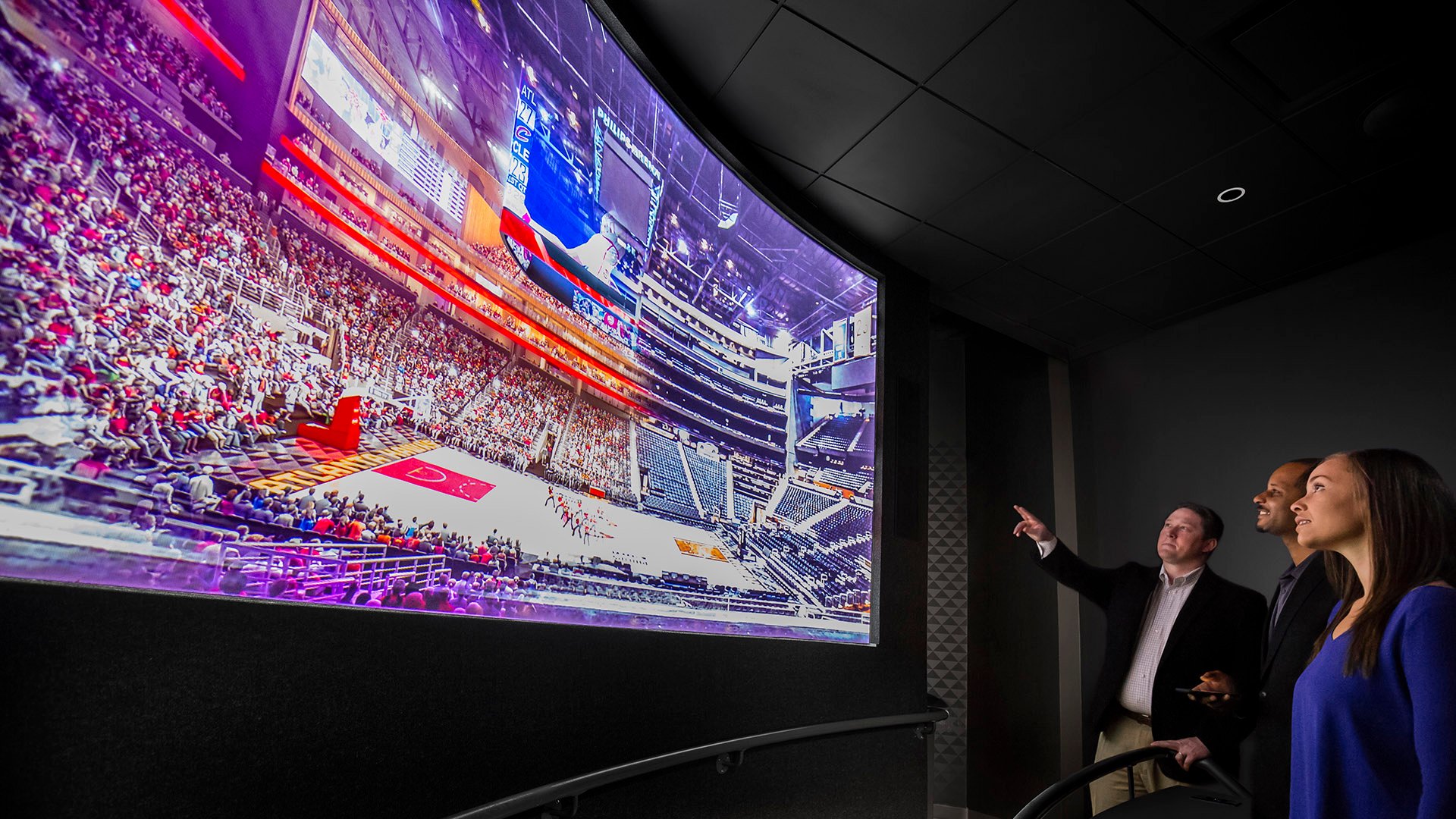 Three individuals stand in a sleek, modern exhibit space, captivated by a massive curved screen displaying a high-resolution image of a vibrant basketball arena packed with fans. One person points toward the screen, engaging the group in discussion. The immersive display simulates the atmosphere of a live sporting event, likely designed to explore sports culture, stadium experiences, or fan engagement in a museum or entertainment venue.