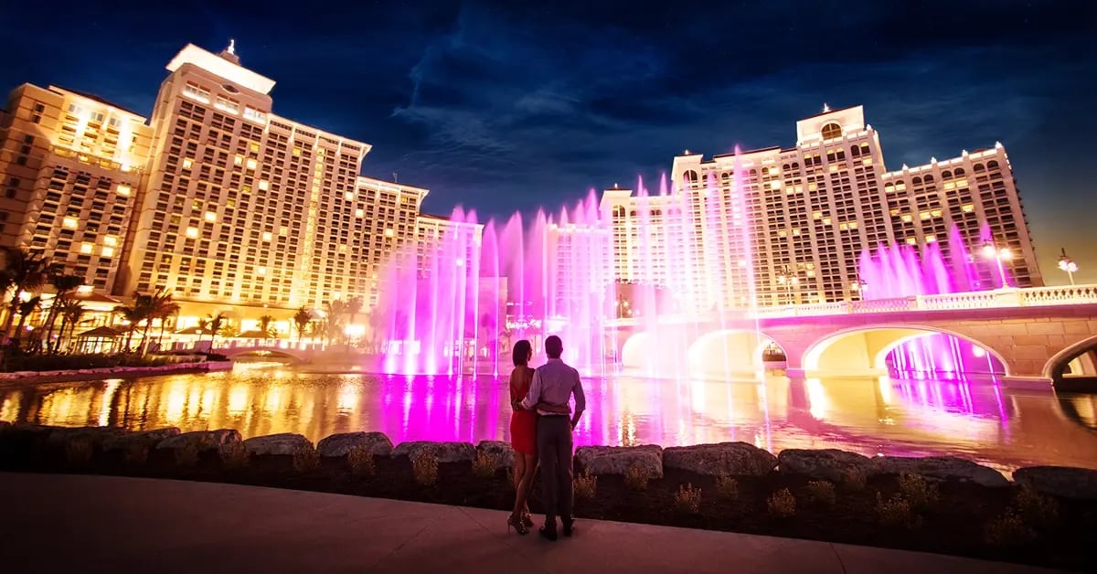 Two guests stand outside at a resort, watching a nighttime fountain show illuminated with vibrant, choreographed lighting effects.