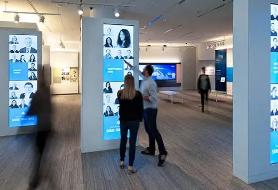 Two visitors to the Cox Discovery Center interacting with a large touchscreen totem in order to learn more about the employees that call Cox home.
