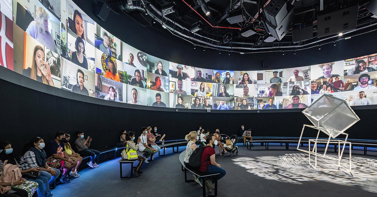 Visitors sit on curved benches in a dark, circular room with a panoramic screen displaying a large grid of video call participants.