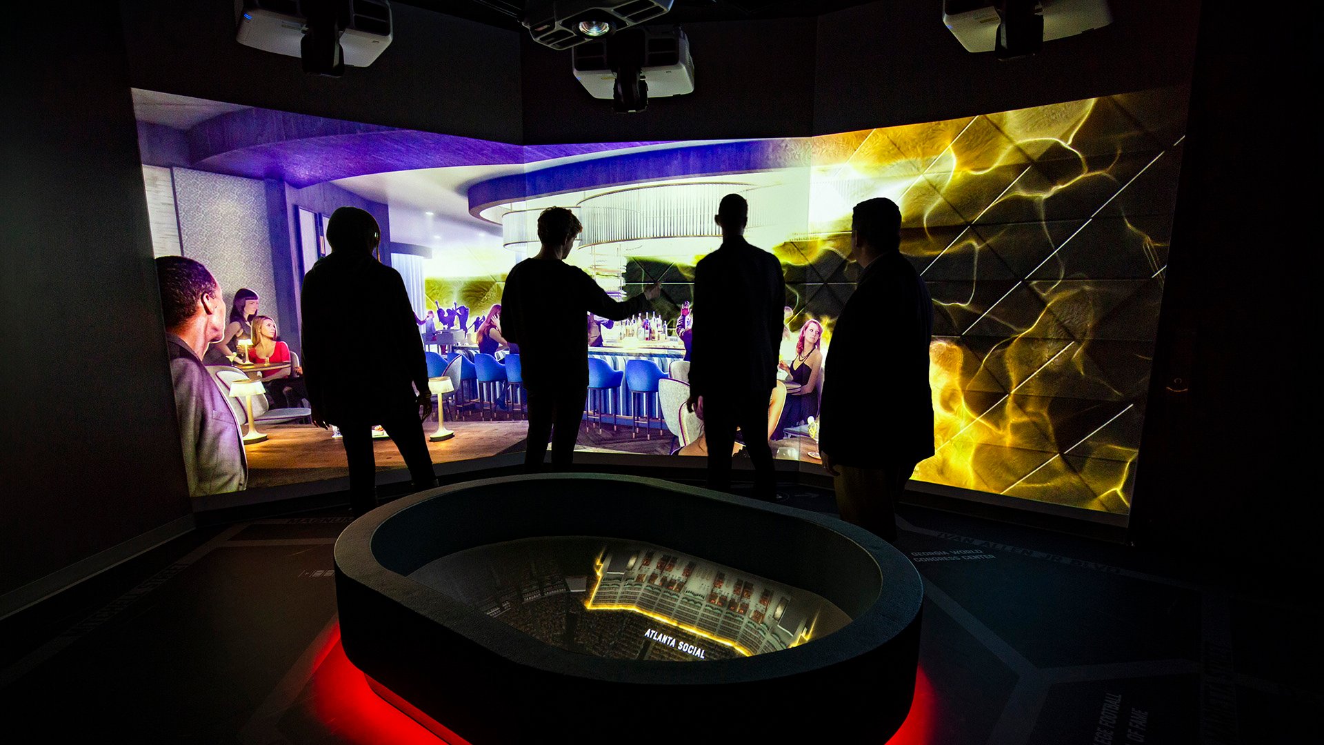 Visitors stand in a dark, immersive exhibit room featuring a central oval display with glowing red lights at its base. Surrounding the visitors is a vibrant, wall-sized video projection depicting a lively social scene in a modern bar or lounge setting. The exhibit creates an engaging, interactive environment that likely explores themes of culture, communication, or social dynamics through multimedia storytelling.