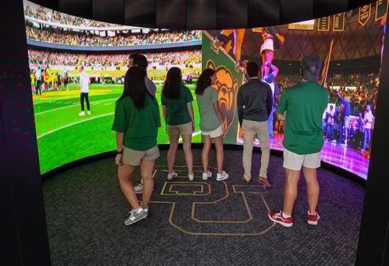 Visitors stand inside an immersive curved screen theater showcasing dynamic Baylor University sports moments, creating an engaging and interactive fan experience.